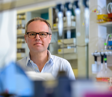Gero Miesenböck, a white man with thin brown grey hair and glasses. He stands in a laboratory with out of focus pipettes in the foreground.