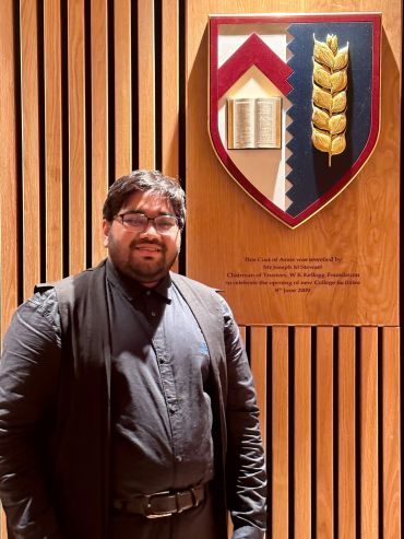 Taqi stands smiling in front of a wooden wall featuring the Kellogg College coat of arms.