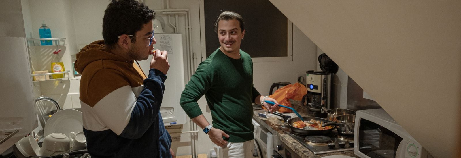 Two boys stand in a student kitchen. One is leaning against the counter whilst the other is cooking various things in a frying pan on the stove.