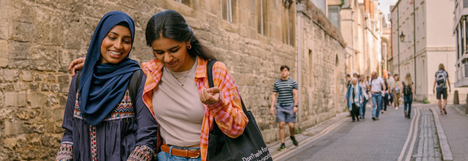 A photograph of two girls walking down a street in Oxford together. One girl has her arm around the other, and they are both smiling and laughing whilst they walk.