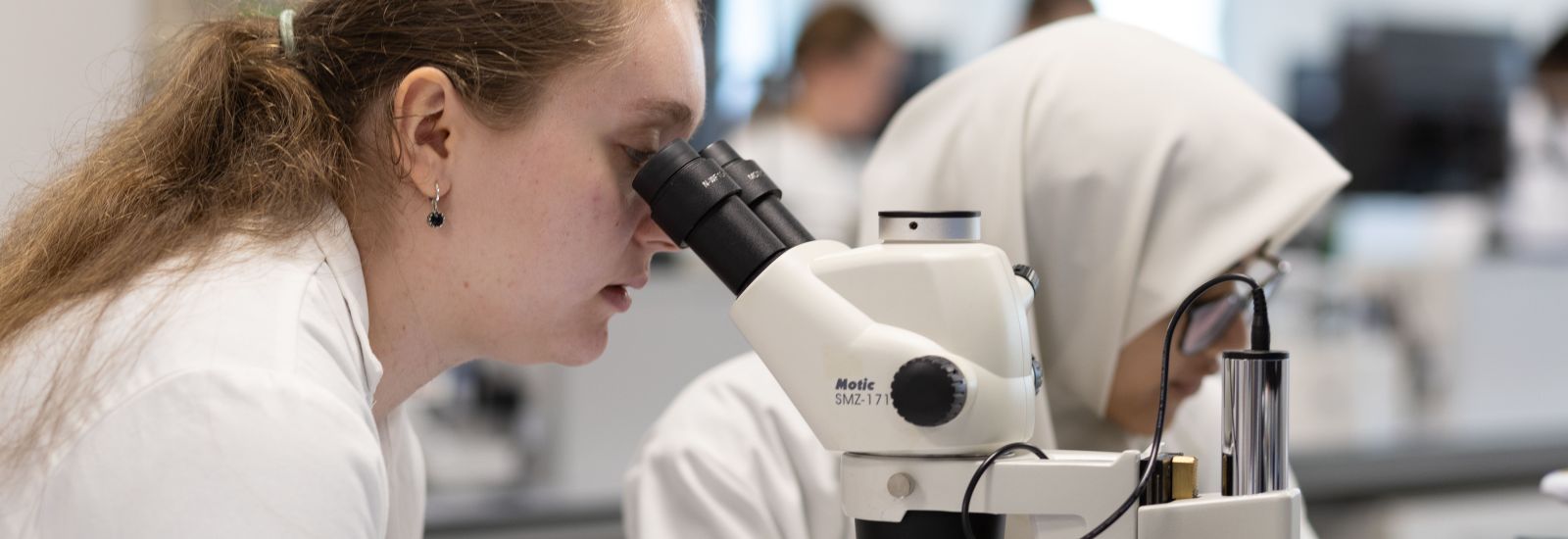 A girl in a lab coat looks through the lenses on a microscope in one of Oxford University's labs.