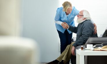 A female caregiver helps a senior man to get up from a sofa.